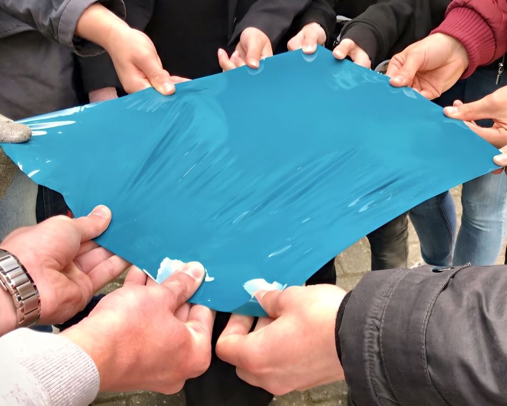 Handen van studenten middelbaar beroepsonderwijs houden samen stuk zelfklevende folie vast tijdens Appcademy gastcollege op ROC / MBO opleiding signmaker