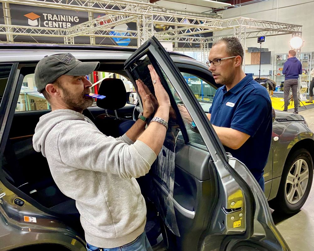 Cursist doet praktijkoefening raamfolie aanbrengen op zijruit met draairaam met toekijkende trainer tijdens Appcademy Automotive Windowfilm cursus