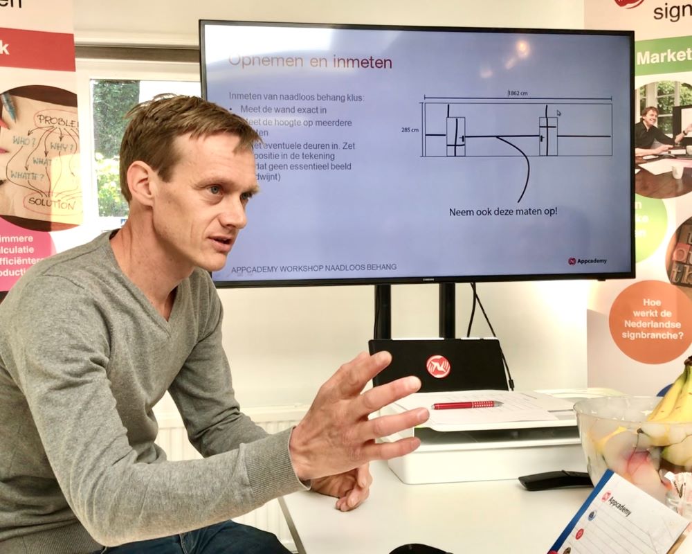 Trainer geeft uitleg over inmeten van wanden m.b.v. power point via LCD scherm tijdens Appcademy Naadloos fotobehang cursus