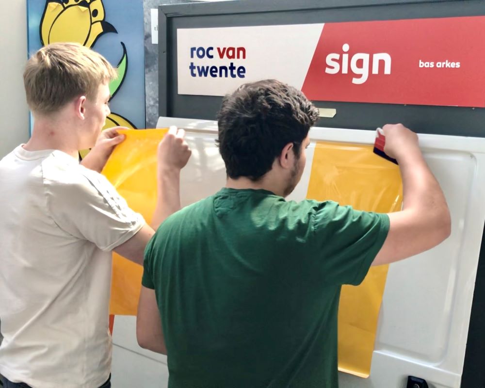 2 Studenten doen praktijkoefeningen folie plakken met rakeltechniek tijdens Appcademy workshop en gastcollege op ROC / MBO signopleiding