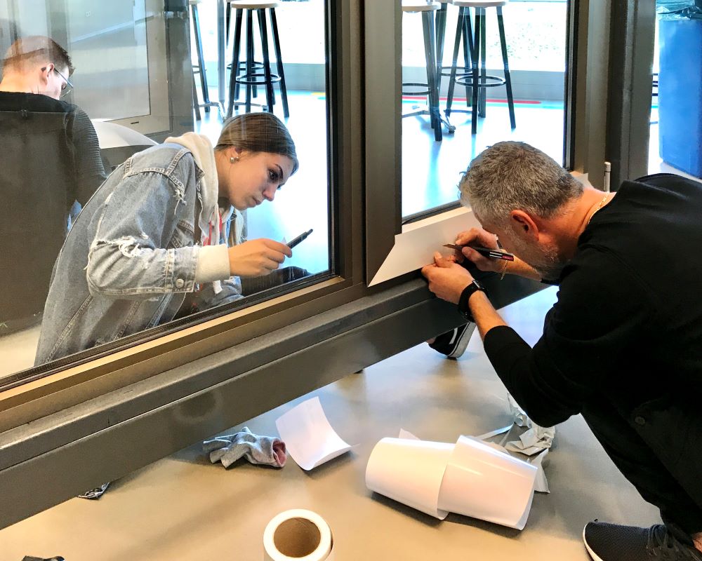 Twee cursisten doen zittend praktijkoefeningen op aluminium kozijn tijdens “Schilderen met folie” Facadefilm cursus